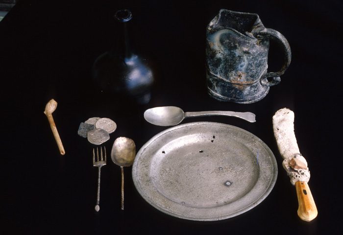 Caption 28- Place setting fit for a Captain. Silver plate, silver fork and spoons and probably a bone handled knife. Near the fork is a small group of silver reales. Note the tankard and onion bottle which might have held wine. The dinner is concluded with a smoke from a clay pipe. -1715 Fleet
