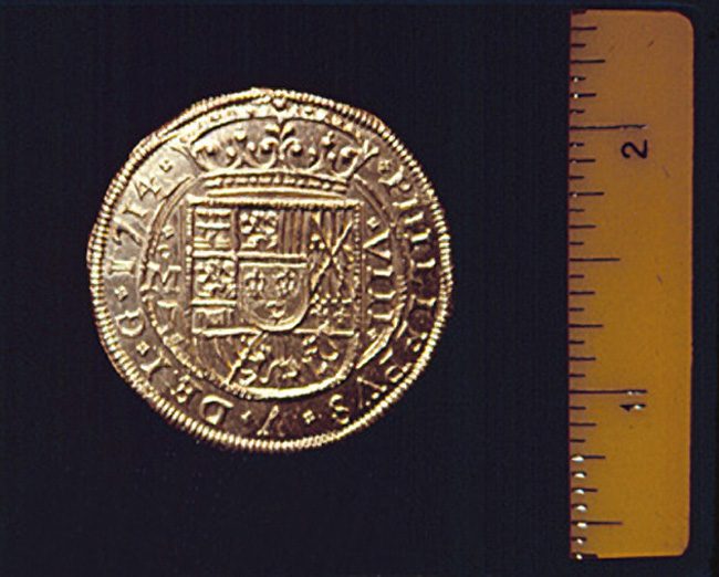 Spain's imperial grandeur imprints a golden doubloon. About the size of a silver dollar, it is here enlarged to show markings. "Phillip V, By the Grace of God, 1714," reads the outer ring of lettering. "M" at left signifies minting in Mexico City. "J" is the mark of the assayer, Jose Eustaquio de Leon. "VIII" tells the value, eight escudos.  The shield combined coats of arms of Spain's provinces and possessions. Fleur-de-lis of the Bourbons, the King's family, fills the center. Castle tower in upper left symbolizes Castile. Rampant lion at top represents Leon. Followed at the bottom by the Low Countries, and Spanish Netherlands. Vertical bars at top right denote Argon. Elongated bars with cross beams stand for Naples and Italy.